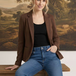 A woman wearing a graced brown knit blazer over a black top and blue jeans, sitting on a wooden bench.