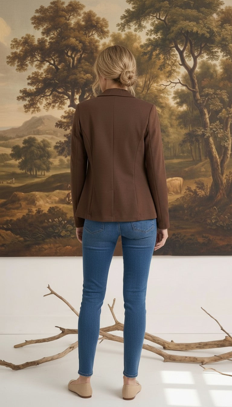 Woman wearing a brown graced knit blazer jacket with blue jeans, viewed from the back against a landscape painting.