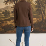 Woman wearing a brown graced knit blazer jacket with blue jeans, viewed from the back against a landscape painting.