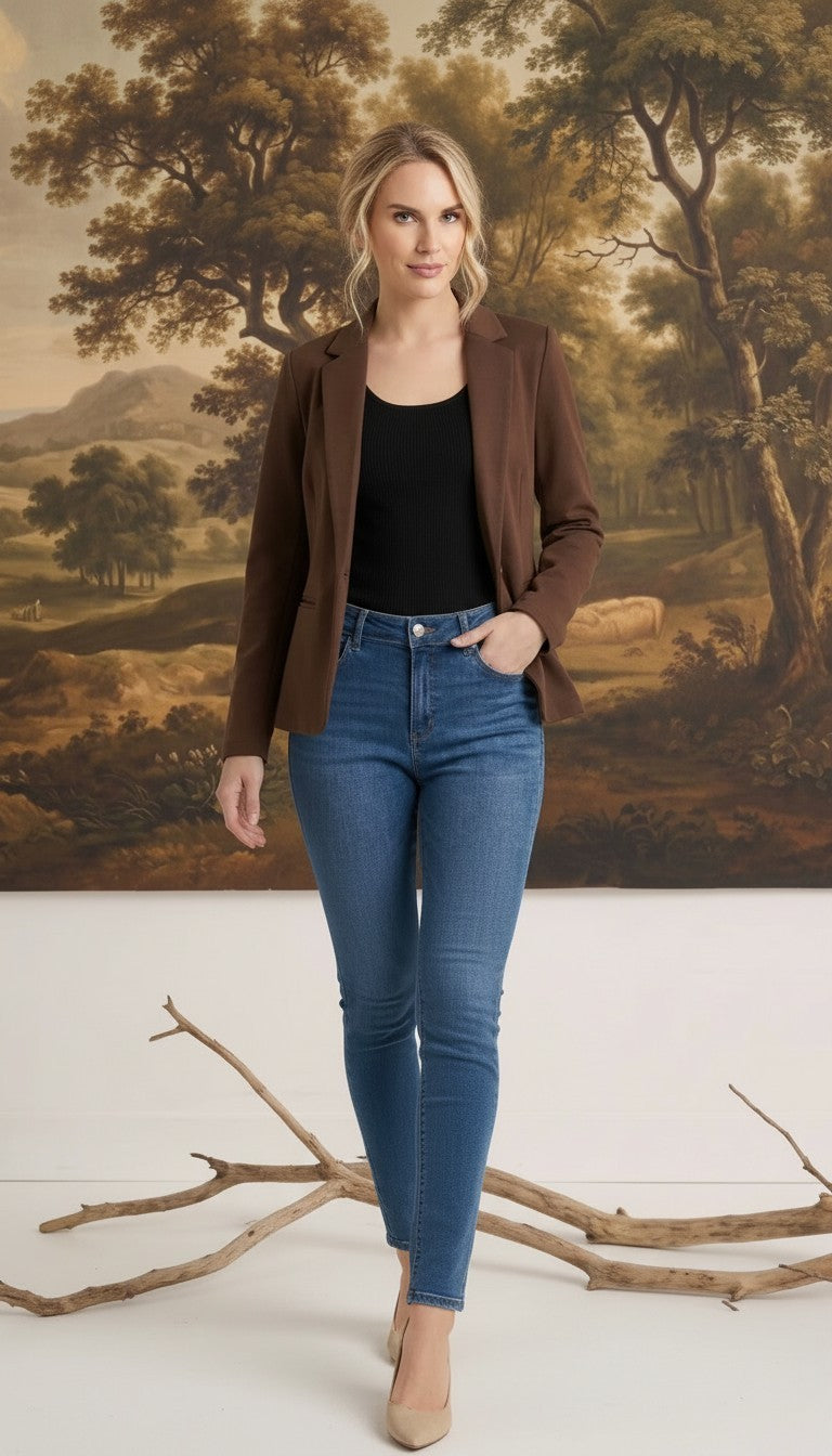 A woman wearing a graced brown knit blazer over a black top and blue jeans, standing in front of a landscape painting.