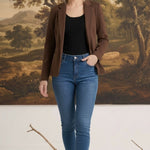 A woman wearing a graced brown knit blazer over a black top and blue jeans, standing in front of a landscape painting.