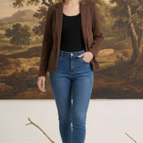A woman wearing a graced brown knit blazer over a black top and blue jeans, standing in front of a landscape painting.
