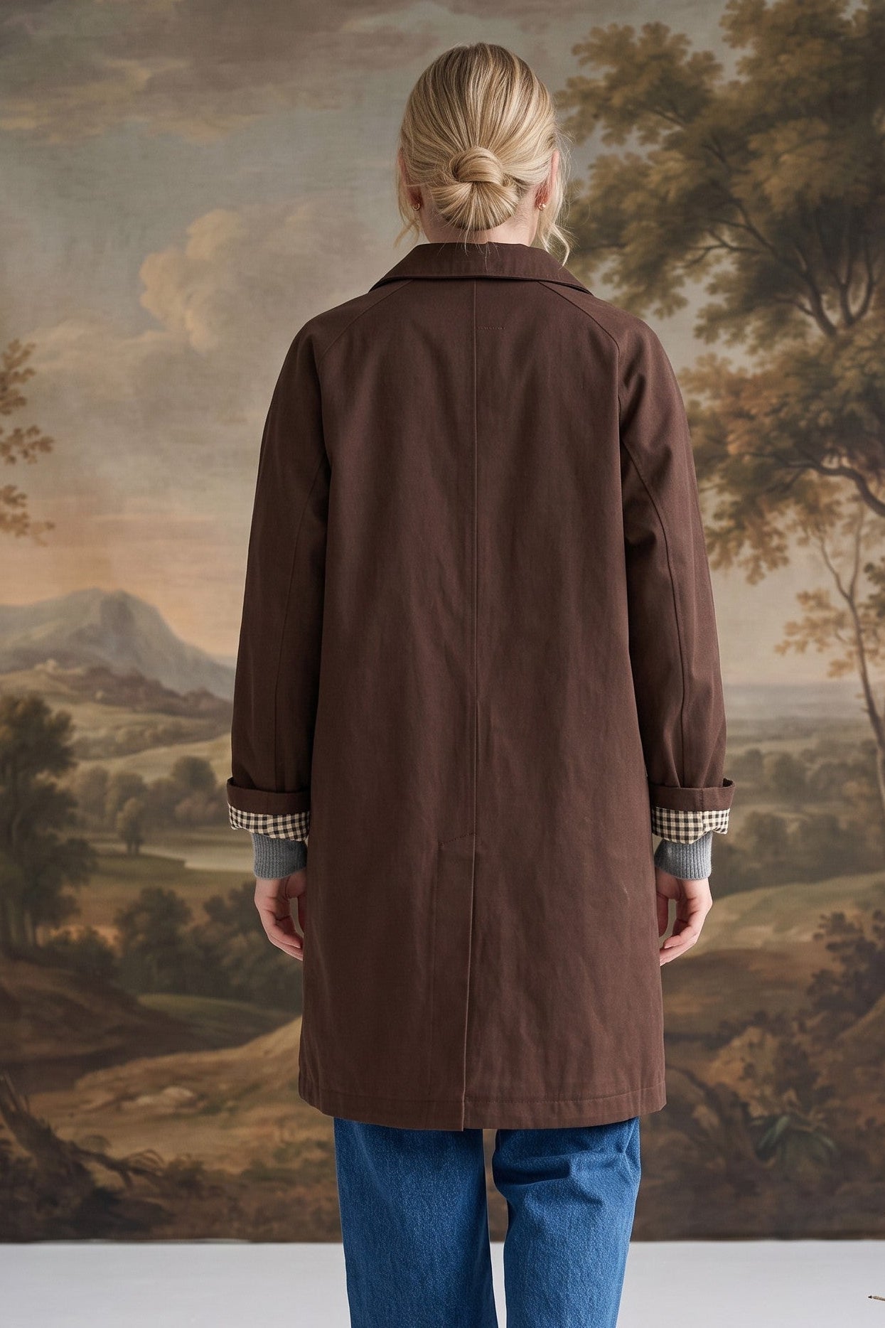 Back view of a woman wearing a graced brown midi trench coat with cuffed sleeves revealing a gingham lining