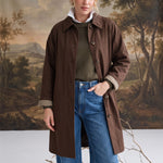 Model wearing a graced brown midi trench coat with a classic collar and plaid lining over jeans and a green sweater.