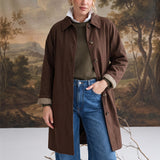 Model wearing a graced brown midi trench coat with a classic collar and plaid lining over jeans and a green sweater.