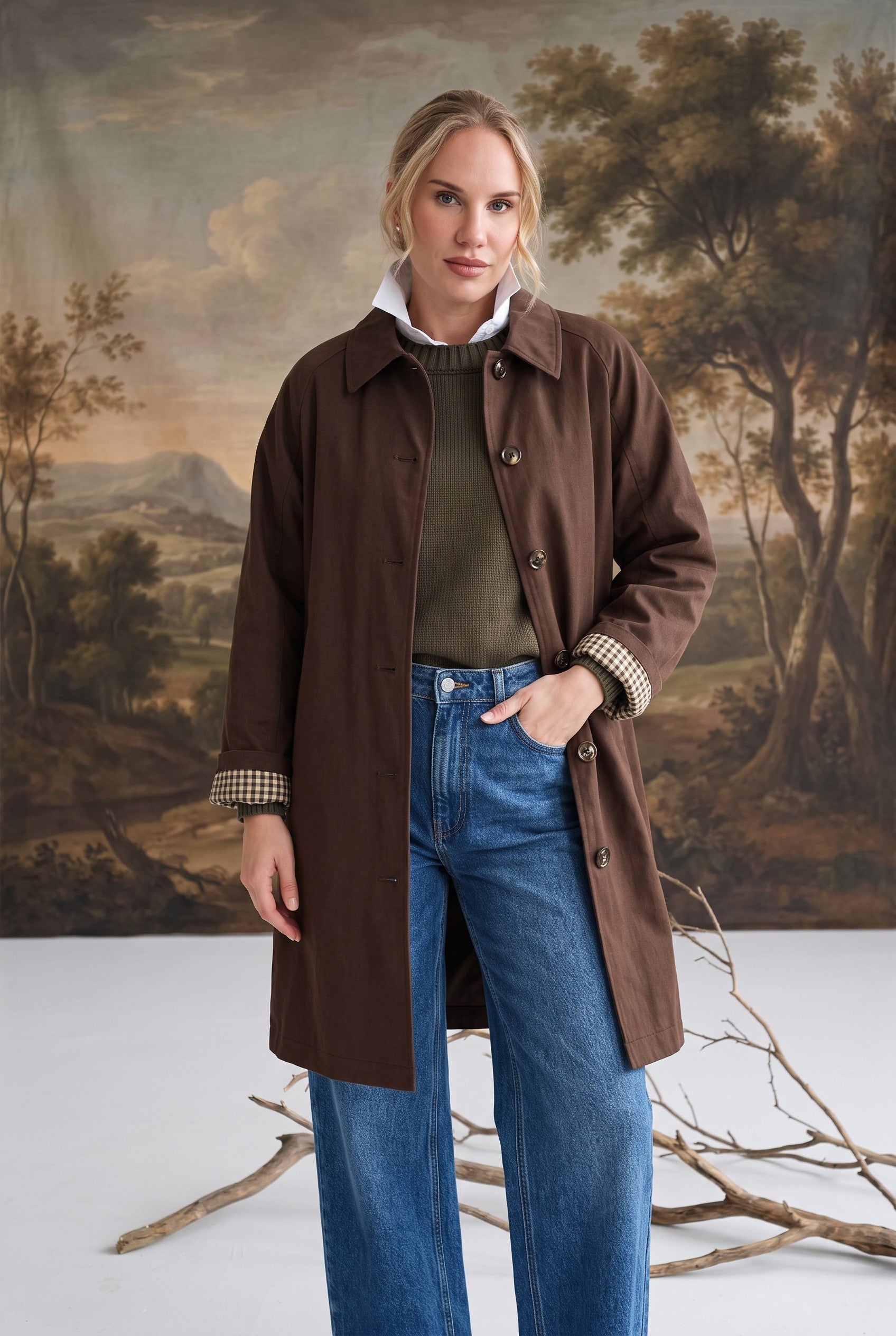 Model wearing a graced brown midi trench coat with a classic collar and plaid lining over jeans and a green sweater.
