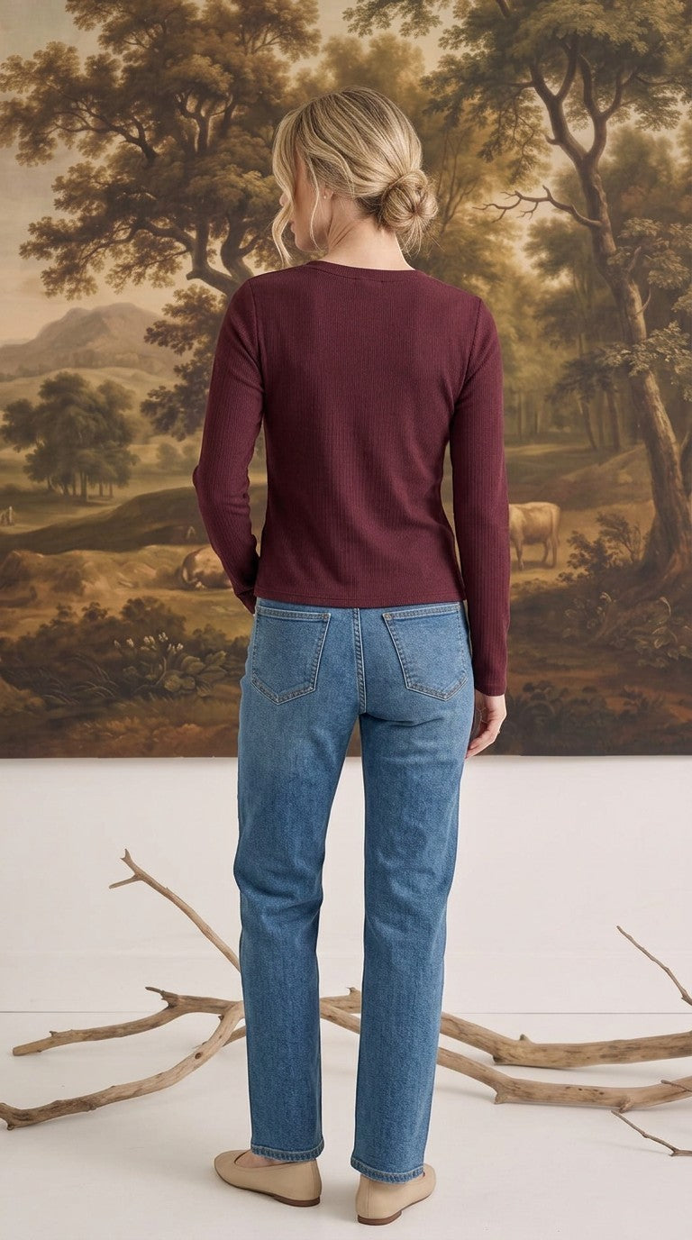 A woman wearing a graced burgundy ribbed long sleeve top and blue jeans, viewed from the back in a studio setting.