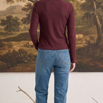 A woman wearing a graced burgundy ribbed long sleeve top and blue jeans, viewed from the back in a studio setting.