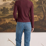 A woman wearing a graced burgundy ribbed long sleeve top and blue jeans, viewed from the back in a studio setting.