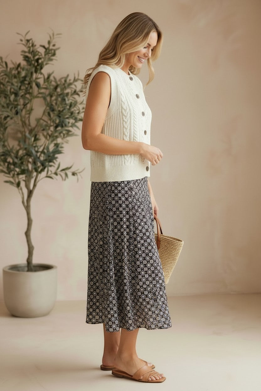 Model wearing a cream graced cable knit sweater vest with buttons, paired with a patterned midi skirt and sandals.