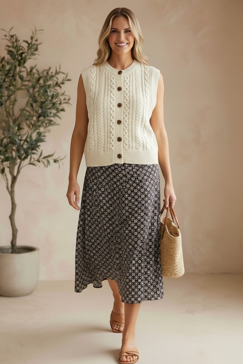 Model wearing a cream graced cable knit button-up sweater vest with a patterned midi skirt and straw bag.