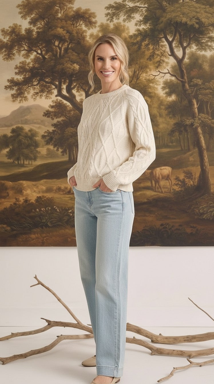 Woman wearing a cream graced crew neck cable knit sweater with light wash jeans in front of a landscape painting