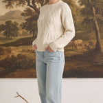 Woman wearing a cream graced crew neck cable knit sweater with light wash jeans in front of a landscape painting