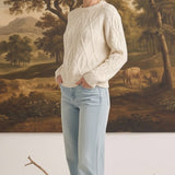 Woman wearing a cream graced crew neck cable knit sweater with light wash jeans in front of a landscape painting