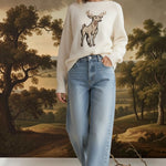 Model wearing a cream graced deer graphic knit sweater with light wash denim jeans in a neutral indoor setting.