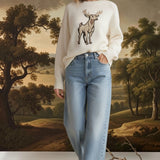 Model wearing a cream graced deer graphic knit sweater with light wash denim jeans in a neutral indoor setting.