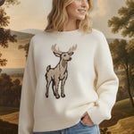 A woman wearing a cream graced deer graphic sweater with blue jeans in a sunlit room.
