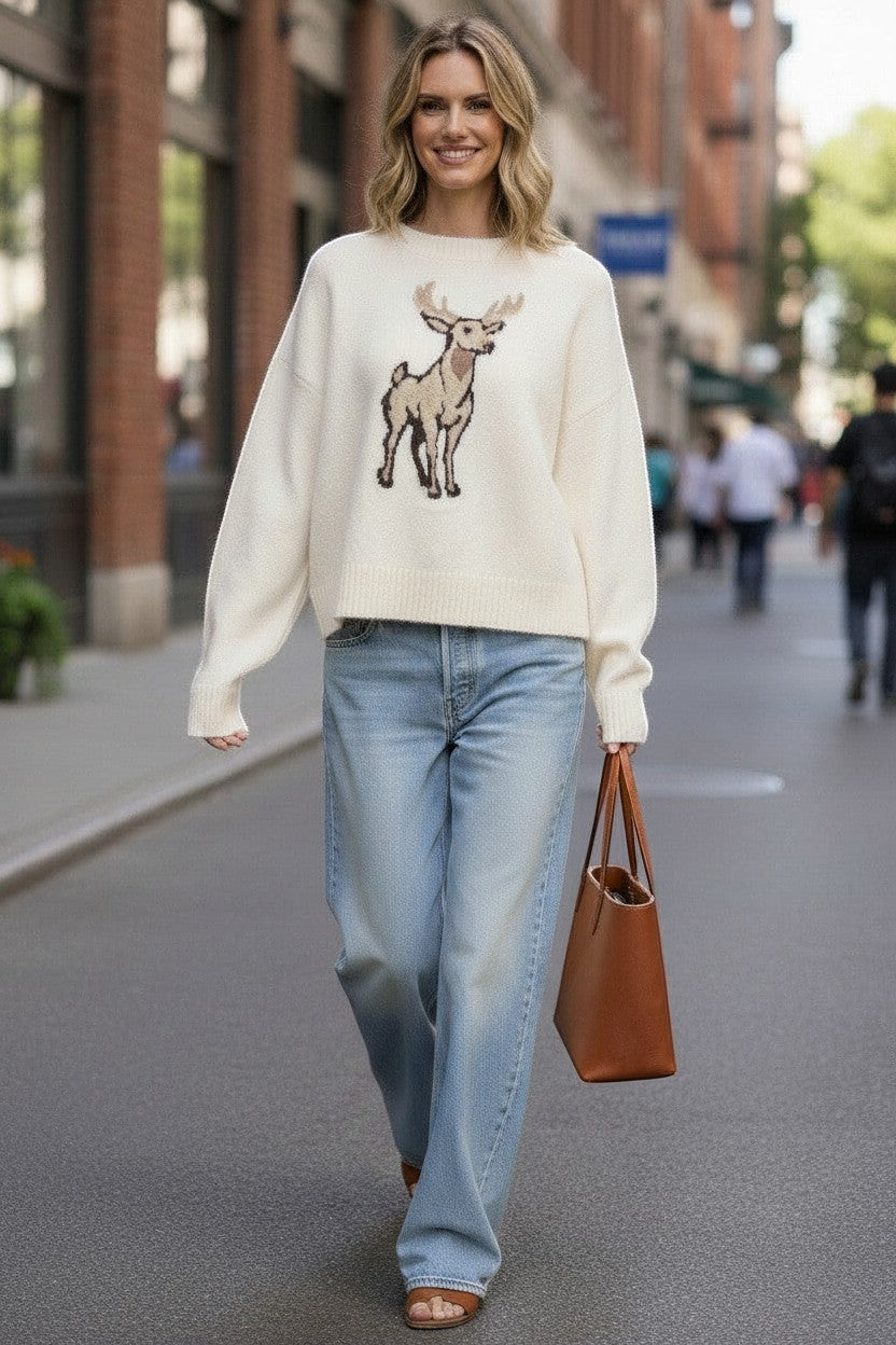 A woman wearing a graced cream-colored knit sweater with a deer graphic, paired with light-wash jeans and a tote bag.