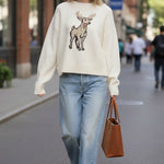 A woman wearing a graced cream-colored knit sweater with a deer graphic, paired with light-wash jeans and a tote bag.