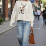 A woman wearing a graced cream-colored knit sweater with a deer graphic, paired with light-wash jeans and a tote bag.