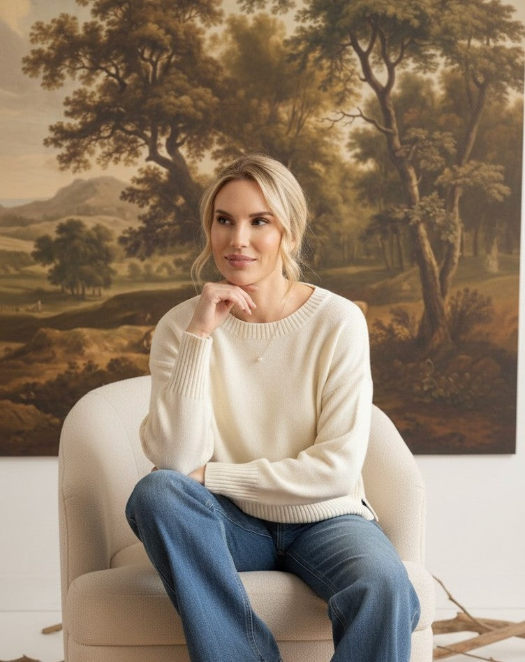 A woman wearing a graced ivory Eloise crew neck sweater with dropped shoulders, sitting in a chair against a landscape painting.