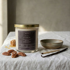 Candle with a label on a table with vanilla beans and a bowl in a softly lit room.