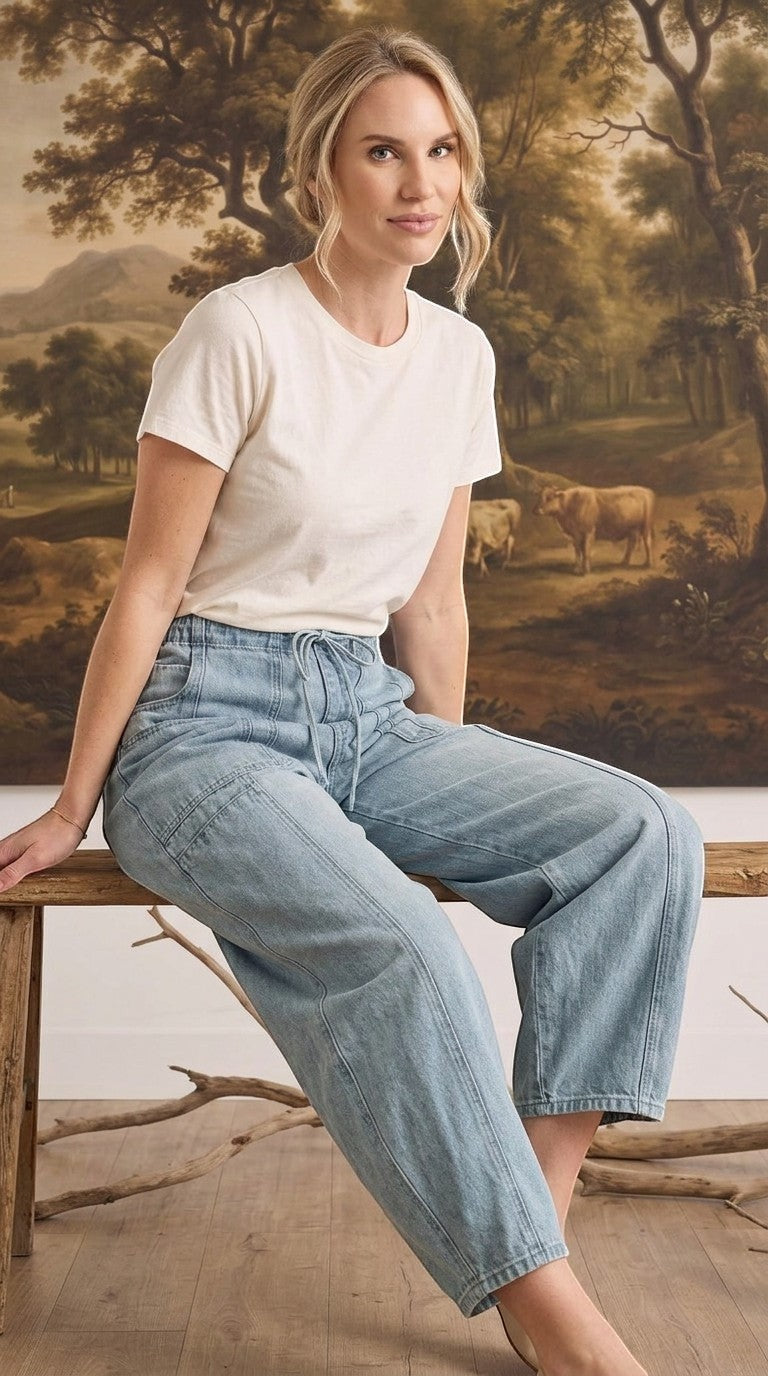 Model wearing graced light wash soft denim barrel pants with a white t-shirt, sitting on a rustic wooden bench.
