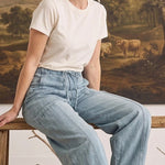Model wearing graced light wash soft denim barrel pants with a white t-shirt, sitting on a rustic wooden bench.