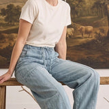 Model wearing graced light wash soft denim barrel pants with a white t-shirt, sitting on a rustic wooden bench.