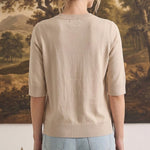 Back view of a woman wearing a graced oatmeal short sleeve knit top with a ribbed crew neck and hem