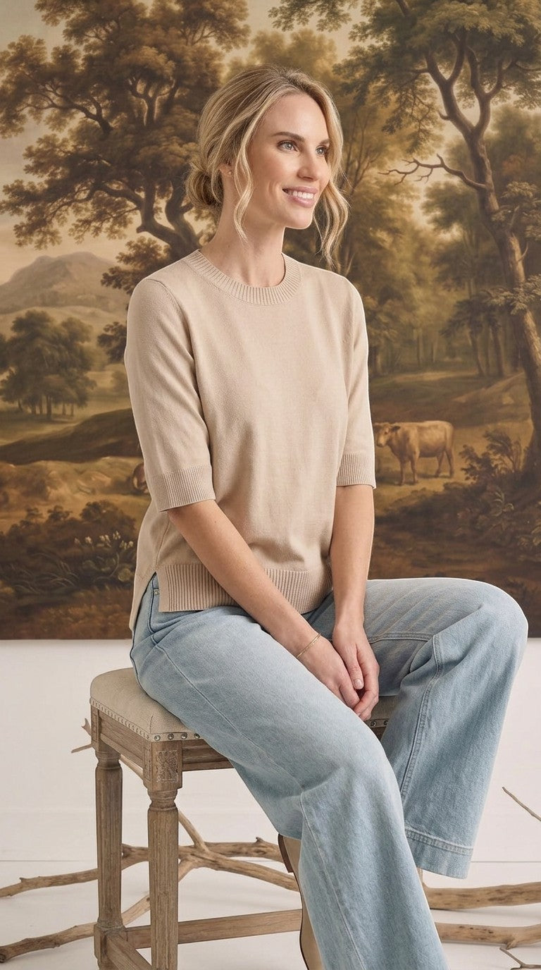 A model wearing a graced oatmeal short sleeve knit top with a crew neckline and ribbed trim, paired with light jeans.