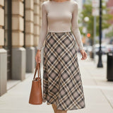 A model wearing a graced A-line midi skirt with a beige and black plaid pattern, paired with a long-sleeve top.