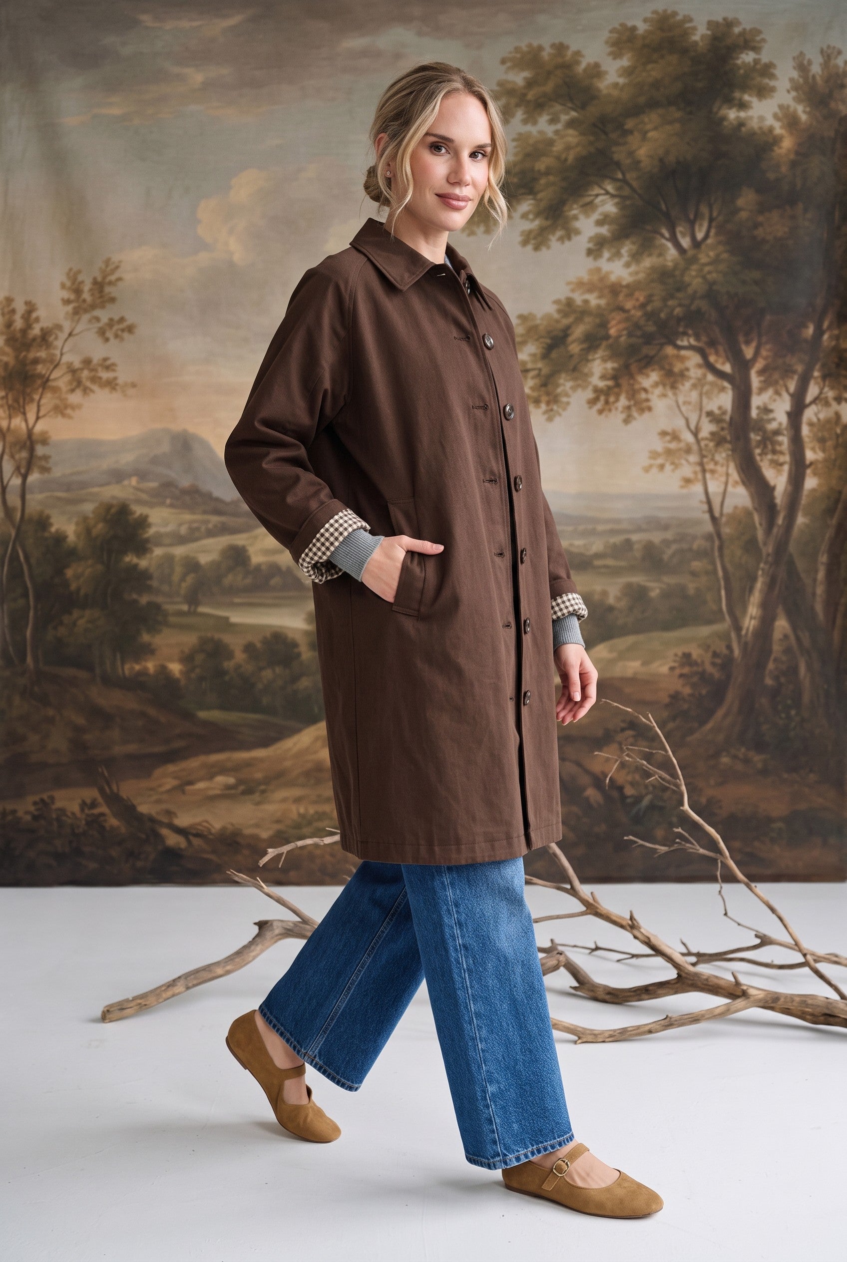 graced The Raven midi trench coat in brown with a classic collar and plaid lining, worn by a model with blue jeans.