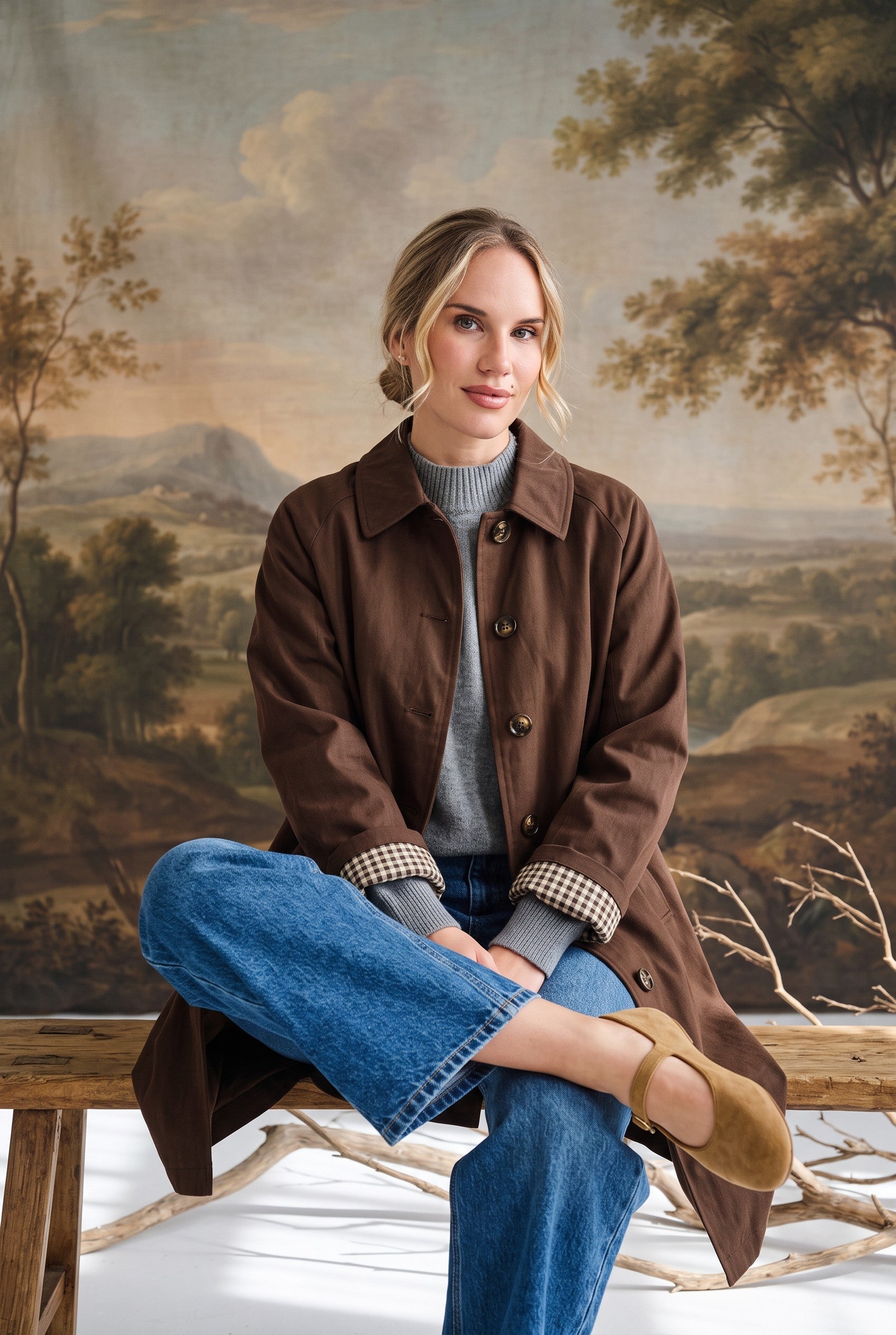 graced The Raven midi trench coat in brown with a classic collar and plaid lining, worn by a woman on a wooden bench.