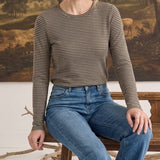 A woman wearing a graced olive and white horizontal striped long sleeve top tucked into blue denim jeans.