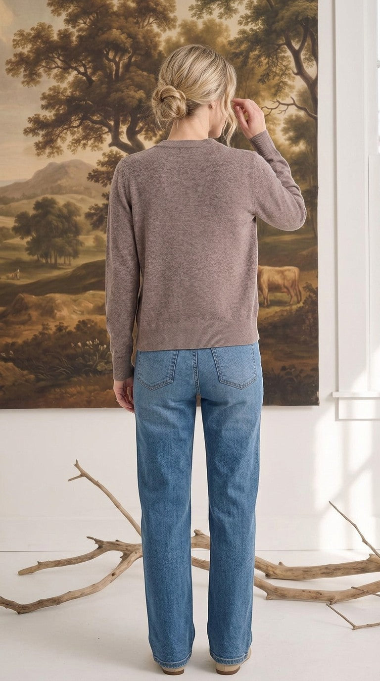 Woman wearing a graced taupe crew neck cardigan with blue jeans, viewed from the back in a rustic studio setting.
