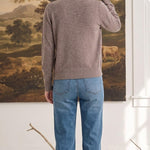 Woman wearing a graced taupe crew neck cardigan with blue jeans, viewed from the back in a rustic studio setting.