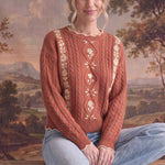 Woman wearing a graced terracotta cable knit sweater with cream floral embroidery and scalloped edges.