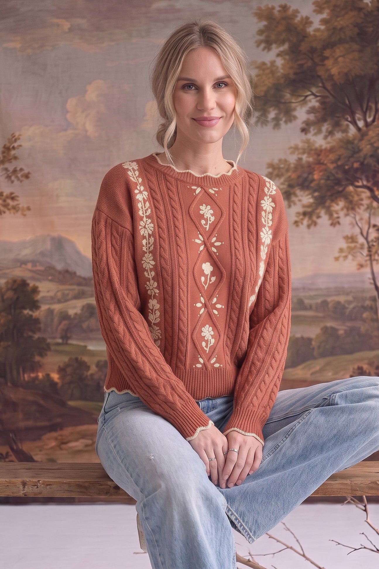 Woman wearing a graced terracotta cable knit sweater with cream floral embroidery and scalloped edges.