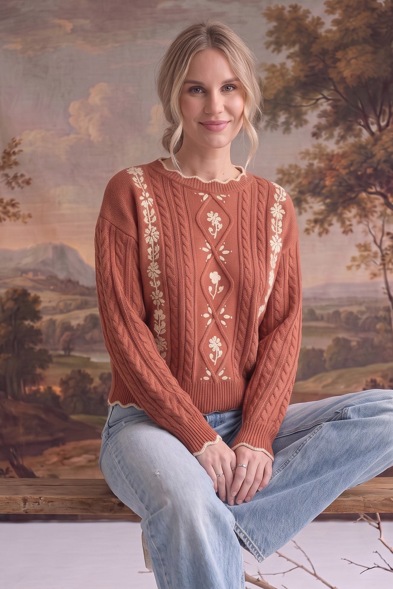 Woman wearing a graced terracotta cable knit sweater with cream floral embroidery and scalloped edges.