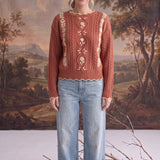 A model wearing a graced terracotta cable knit sweater with delicate floral embroidery and scalloped hem with jeans.