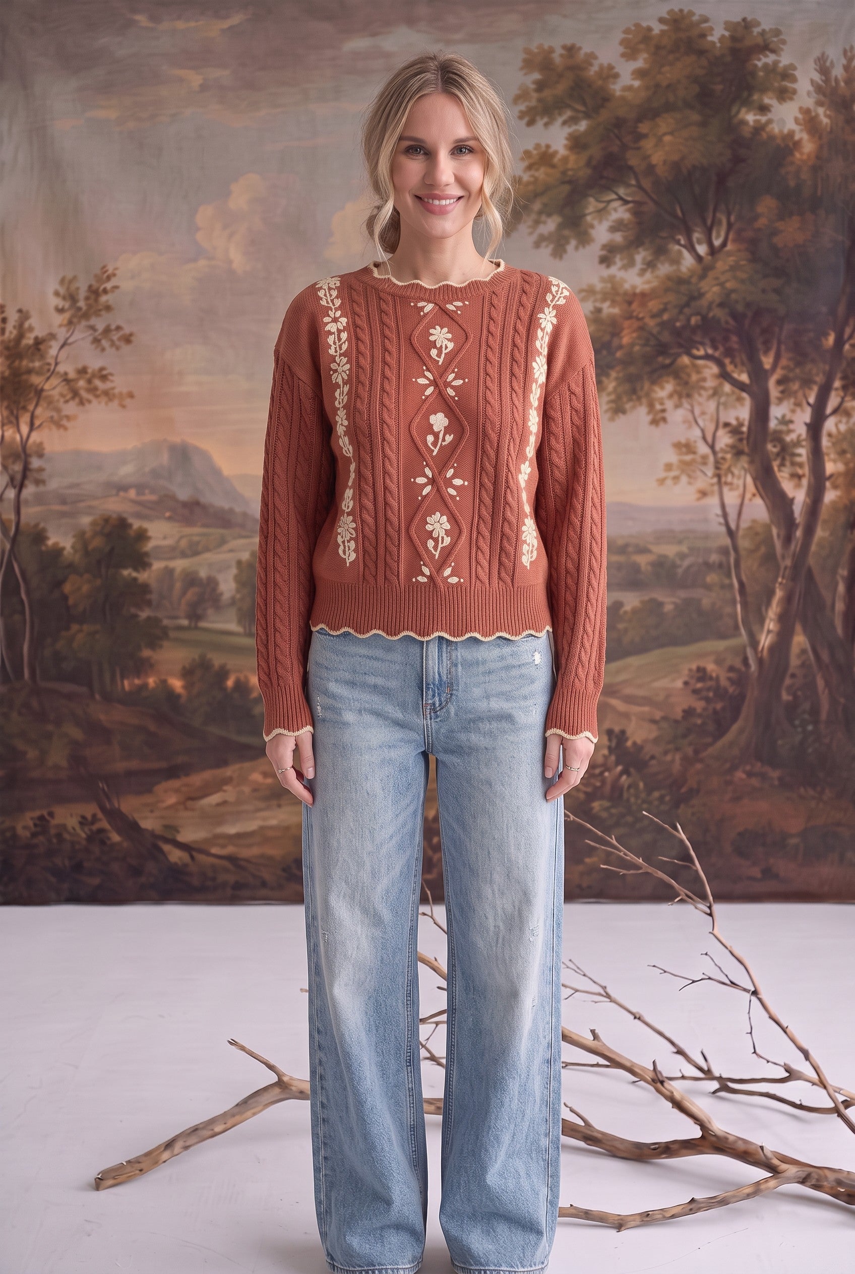 A model wearing a graced terracotta cable knit sweater with delicate floral embroidery and scalloped hem with jeans.
