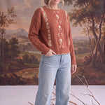 A model wearing a graced terracotta cable knit sweater with delicate floral embroidery and light wash denim jeans.