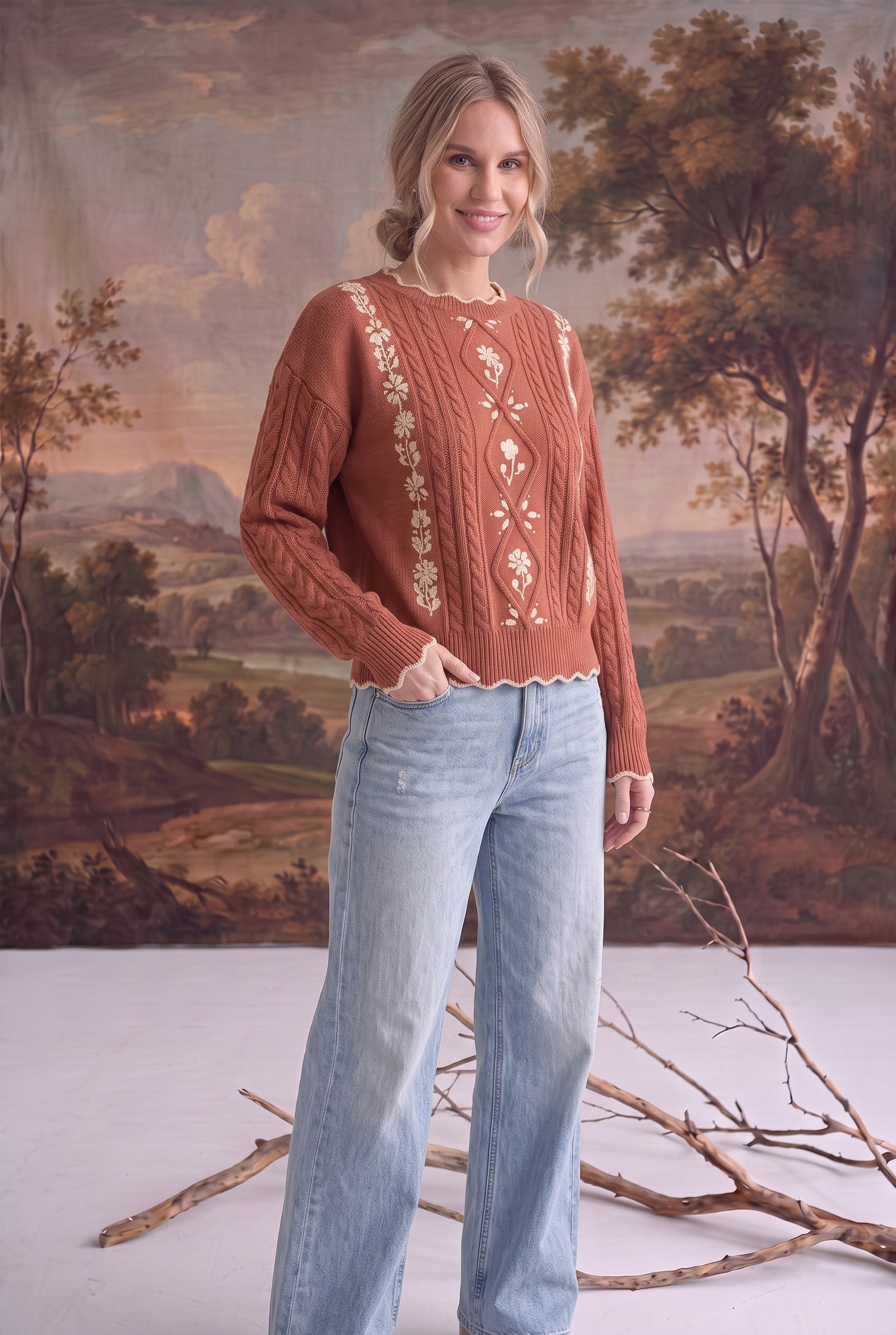 A model wearing a graced terracotta cable knit sweater with delicate floral embroidery and light wash denim jeans.