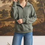 A woman wearing a graced olive green half zip mock neck pullover with long sleeves and blue denim jeans.
