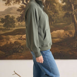 graced olive green half zip mock neck pullover worn by a woman with blue jeans in a studio setting