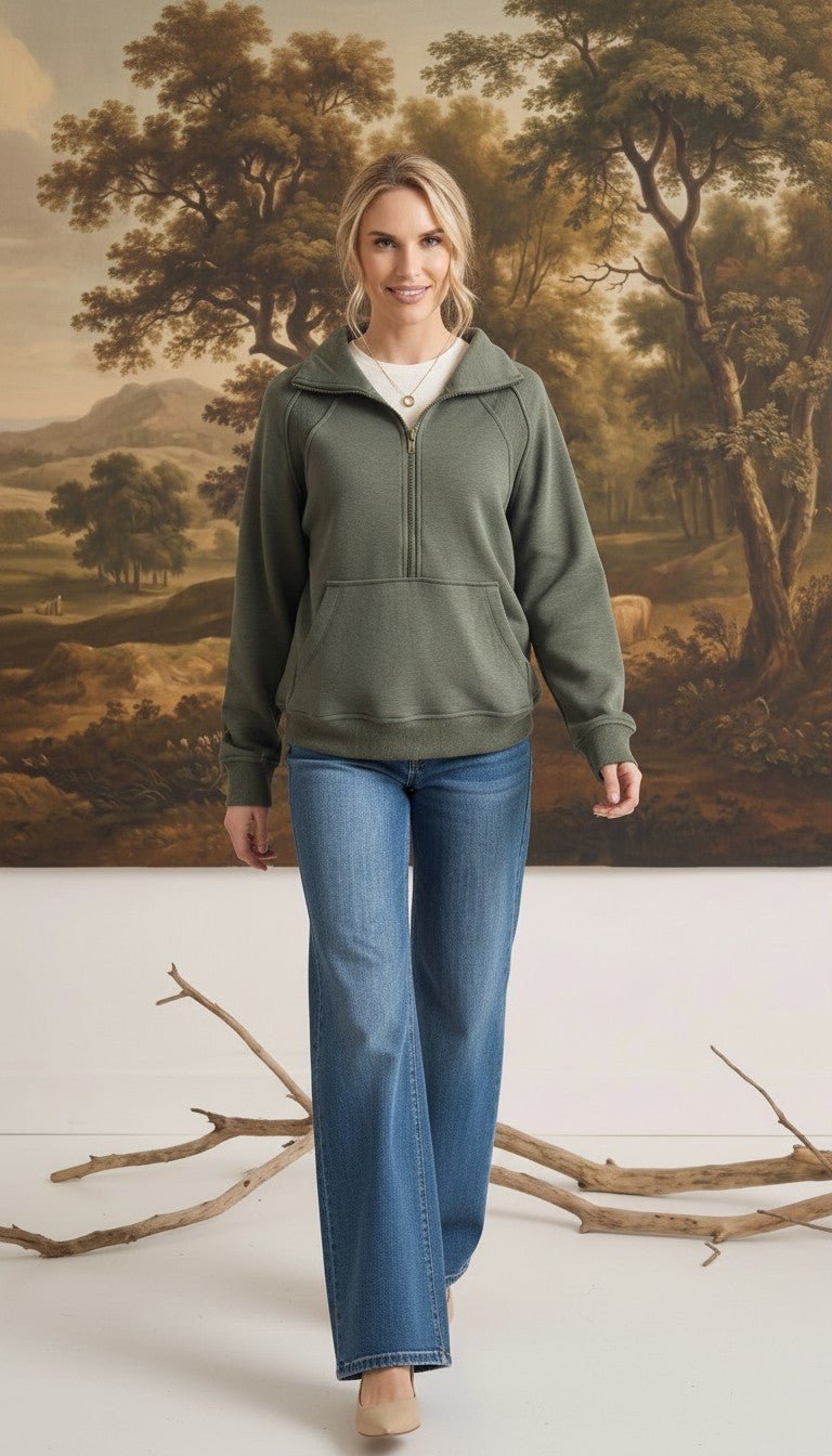 A woman wearing a graced olive green half zip mock neck pullover with blue jeans in a studio setting.