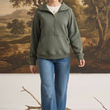 A woman wearing a graced olive green half zip mock neck pullover with blue jeans in a studio setting.