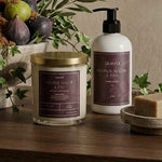 A styled bathroom or spa setup on a wooden surface featuring a jar candle with a gold lid, a white pump bottle with a dark label, a soap bar on a small dish, and a reed diffuser with greenery and figs in the background against a textured beige wall.