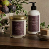 A styled bathroom or spa setup on a wooden surface featuring a jar candle with a gold lid, a white pump bottle with a dark label, a soap bar on a small dish, and a reed diffuser with greenery and figs in the background against a textured beige wall.